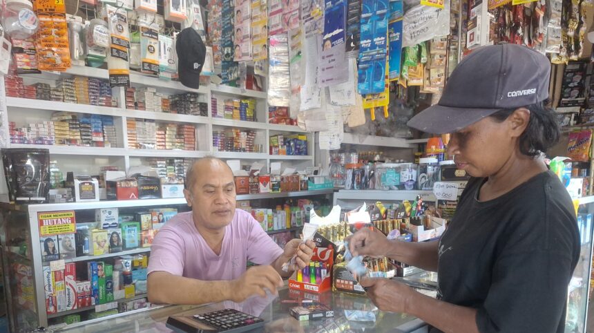 A shop cashier in a pink shirt hands an item to a customer across a glass counter, with shelves full of products behind them.