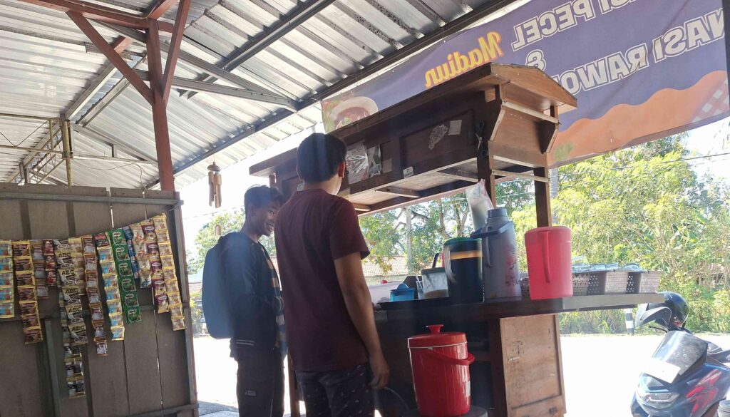 Two young men stand at an open-air snack stand under a metal roof, with packaged snacks hanging on the left and drink dispensers on the counter.