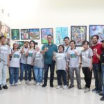 Group of people wearing gray shirts posing in an art gallery in front of colorful paintings on the wall, smiling and making hand gestures.