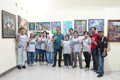 Group of people wearing gray shirts posing in an art gallery in front of colorful paintings on the wall, smiling and making hand gestures.