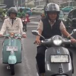 Two scooter riders in traffic: a man wearing a helmet and vest on a gray scooter in the foreground, and a woman in a beige jacket on a mint-green scooter behind him.