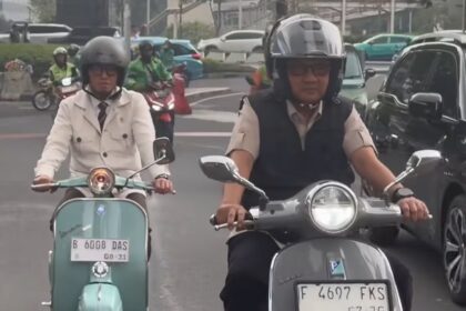 Two scooter riders in traffic: a man wearing a helmet and vest on a gray scooter in the foreground, and a woman in a beige jacket on a mint-green scooter behind him.