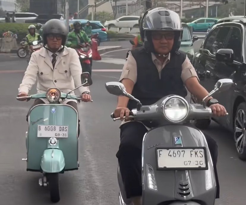 Two scooter riders in traffic: a man wearing a helmet and vest on a gray scooter in the foreground, and a woman in a beige jacket on a mint-green scooter behind him.