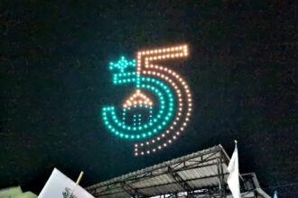 Night scene of a large LED-dot sign forming the number five in teal and orange above a stage at night.