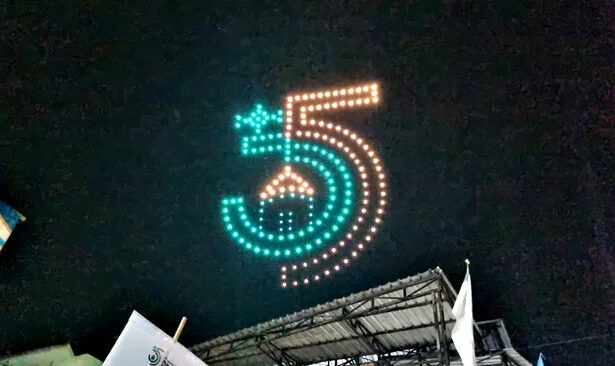 Night scene of a large LED-dot sign forming the number five in teal and orange above a stage at night.