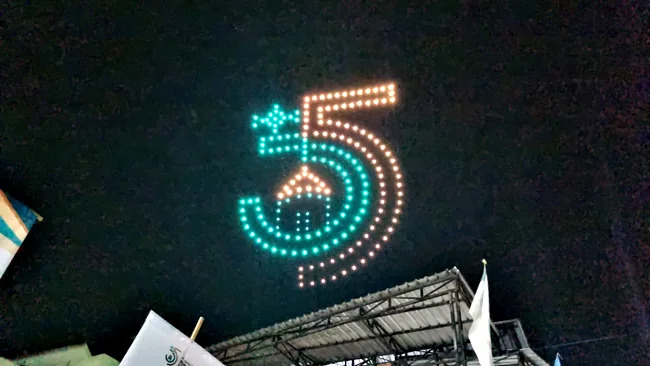 Night scene of a large LED-dot sign forming the number five in teal and orange above a stage at night.