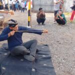 Man in a blue shirt sits on a mat outdoors, practicing archery with a traditional bow while spectators film nearby.