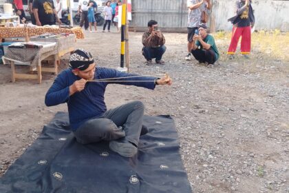 Man in a blue shirt sits on a mat outdoors, practicing archery with a traditional bow while spectators film nearby.