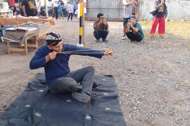 Man in a blue shirt sits on a mat outdoors, practicing archery with a traditional bow while spectators film nearby.