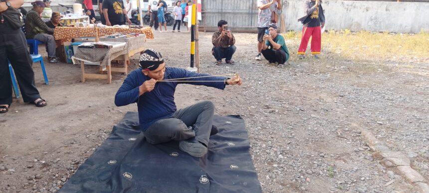 Man in a blue shirt sits on a mat outdoors, practicing archery with a traditional bow while spectators film nearby.