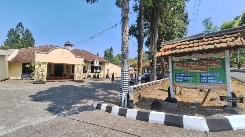 20260916_100917 - Mabur.co Public building with an arched facade and tiled roof, a courtyard, and a large green signboard advertising catering, sound system, and nasi options on the right.
