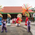 Group of performers in colorful traditional costumes with large masks dancing in an open courtyard under festive paper palm decorations above.