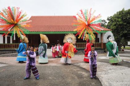Group of performers in colorful traditional costumes with large masks dancing in an open courtyard under festive paper palm decorations above.