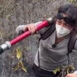 Person wearing a mask and goggles/backpack-belt holding a red fire hose and spraying water through smoky, burnt brush area