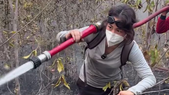 Person wearing a mask and goggles/backpack-belt holding a red fire hose and spraying water through smoky, burnt brush area