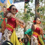 Performers in colorful traditional costumes dancing on a stage outdoors with a decorative backdrop and trees behind them, smiling and moving rhythmically.