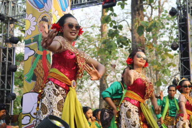 Performers in colorful traditional costumes dancing on a stage outdoors with a decorative backdrop and trees behind them, smiling and moving rhythmically.