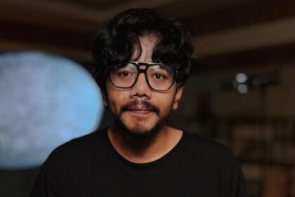Portrait of a man with wavy black hair and a beard, wearing oversized glasses and a black shirt, indoors.