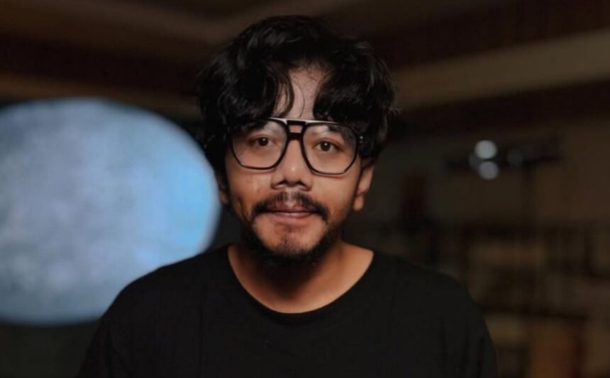 Portrait of a man with wavy black hair and a beard, wearing oversized glasses and a black shirt, indoors.