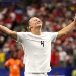 Soccer player in a white jersey with arms raised in celebration on a filled stadium field.