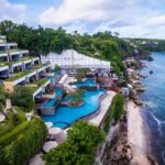 A terraced seaside resort with multiple blue pools, white buildings, and lush greenery along a rocky coastline.