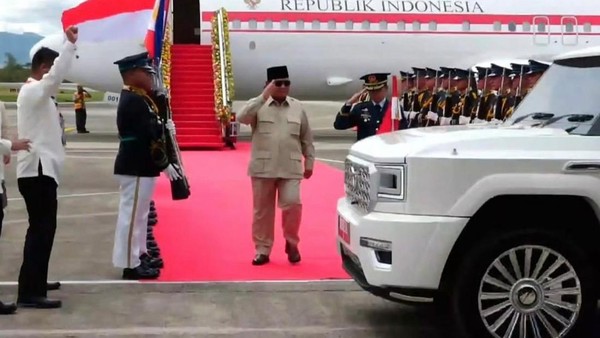Indonesian dignitary walks on a red carpet to greet arrivals, flanked by guards with a white SUV in the foreground.