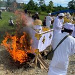 People in white traditional clothing perform a ceremonial fire ritual around a decorated float outdoors. Flames rise from the burning setup as onlookers watch.