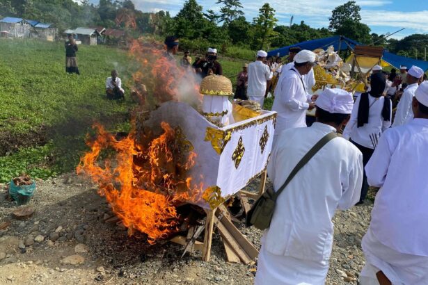 People in white traditional clothing perform a ceremonial fire ritual around a decorated float outdoors. Flames rise from the burning setup as onlookers watch.