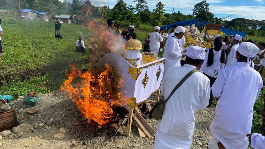 People in white traditional clothing perform a ceremonial fire ritual around a decorated float outdoors. Flames rise from the burning setup as onlookers watch.
