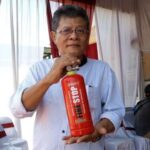 Man wearing glasses and a light striped shirt holds a red fire extinguisher labeled 'Fire Stop' during a demonstration in a room with draped curtains.