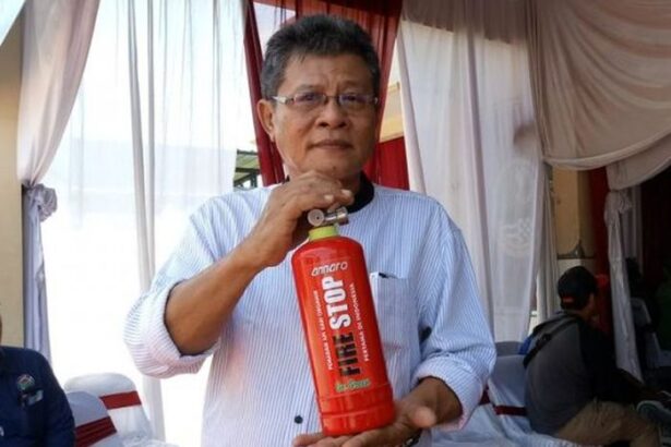 Man wearing glasses and a light striped shirt holds a red fire extinguisher labeled 'Fire Stop' during a demonstration in a room with draped curtains.
