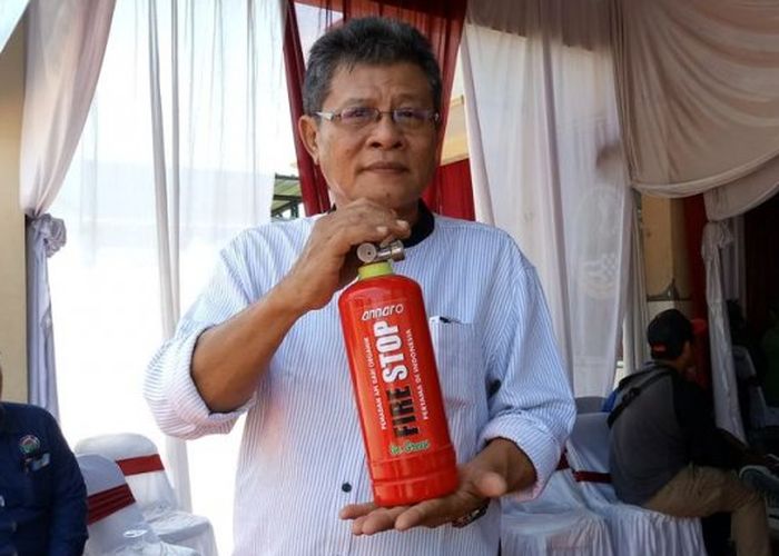 Man wearing glasses and a light striped shirt holds a red fire extinguisher labeled 'Fire Stop' during a demonstration in a room with draped curtains.