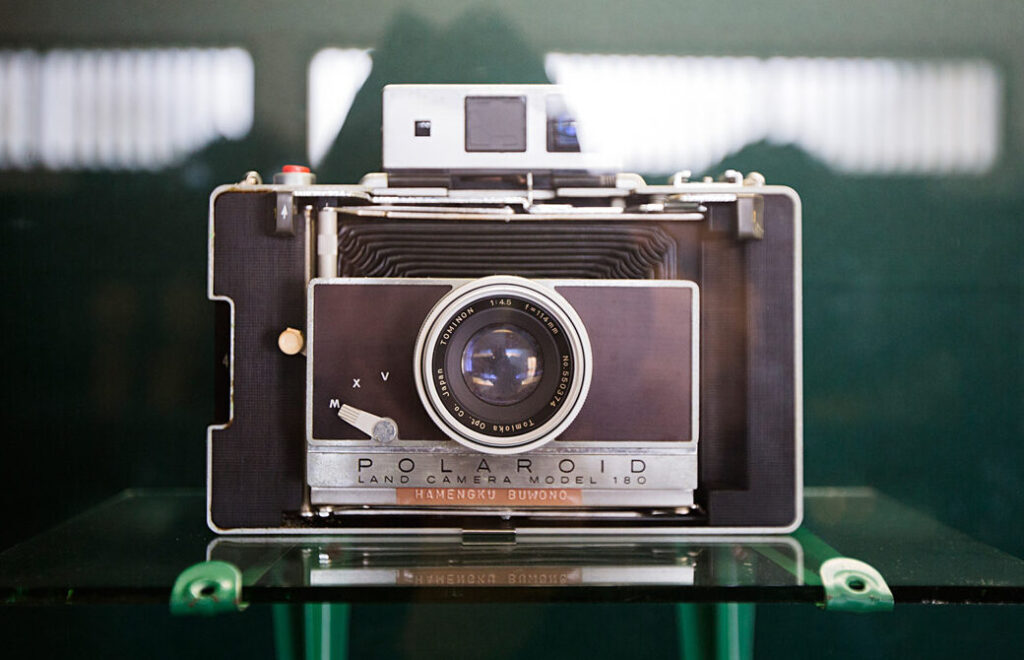 325fc556-24f4-411d-90e4-384183c64ae1 - Mabur.co Polaroid Land Camera Model 180 on a display shelf, front view showing the lens and nameplate.