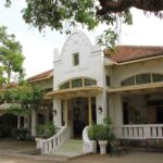 Historic white building with arched windows and a tiled roof, shaded by surrounding trees and a covered veranda.