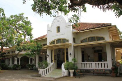 Historic white building with arched windows and a tiled roof, shaded by surrounding trees and a covered veranda.