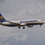 Side view of a Ryanair passenger jet in blue and white livery, with landing gear down, flying against a cloudy sky.