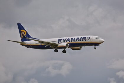 Side view of a Ryanair passenger jet in blue and white livery, with landing gear down, flying against a cloudy sky.