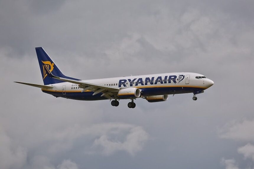 Side view of a Ryanair passenger jet in blue and white livery, with landing gear down, flying against a cloudy sky.