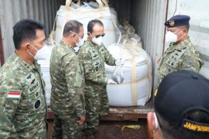 Indonesian soldiers in camouflage and masks inspect large white-wrapped cargo secured inside a shipping container.
