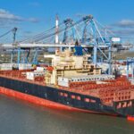 Large cargo ship loaded with colorful shipping containers at a busy port with blue gantry cranes overhead in a clear sky.