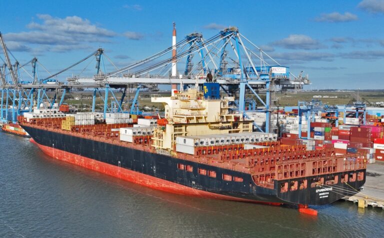 Large cargo ship loaded with colorful shipping containers at a busy port with blue gantry cranes overhead in a clear sky.