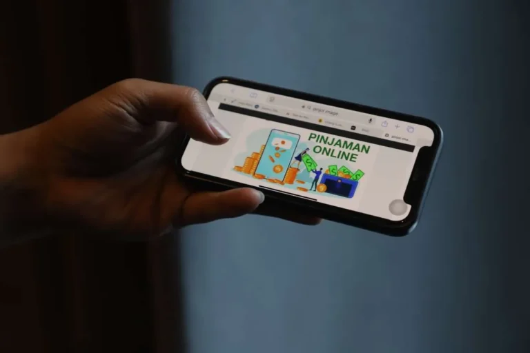 Hand holding a smartphone displaying a graphic with 'PINJAMAN ONLINE' and money icons, suggesting online loans.