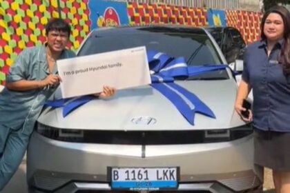 Two people pose beside a silver car with a large blue ribbon; the person on the left holds a sign reading 'I'm a proud Hyundai family'.