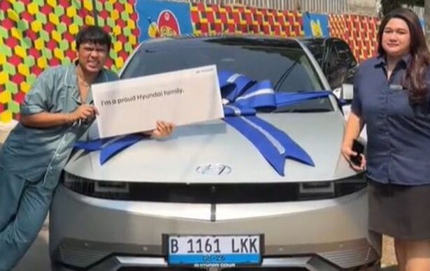 Two people pose beside a silver car with a large blue ribbon; the person on the left holds a sign reading 'I'm a proud Hyundai family'.