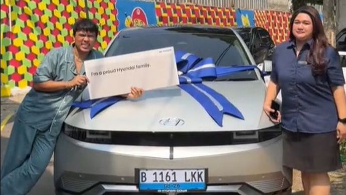 Two people pose beside a silver car with a large blue ribbon; the person on the left holds a sign reading 'I'm a proud Hyundai family'.