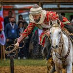 Equestrian performer in a patterned red outfit rides a white horse, leaning forward with a lasso during a crowded event.