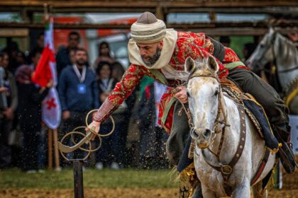 Equestrian performer in a patterned red outfit rides a white horse, leaning forward with a lasso during a crowded event.