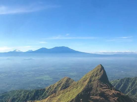 View from a high mountain ridge with jagged peaks, grass in the foreground, and distant blue horizons under a clear sky.