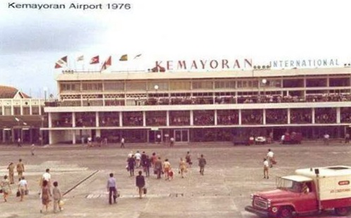 Exterior view of Kemayoran International Airport terminal in 1976, with people walking on the tarmac and a red-and-white bus in the foreground.