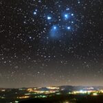 Star-filled night sky over a lit town on rolling hills, with a blue-trim cluster of stars near the top center.